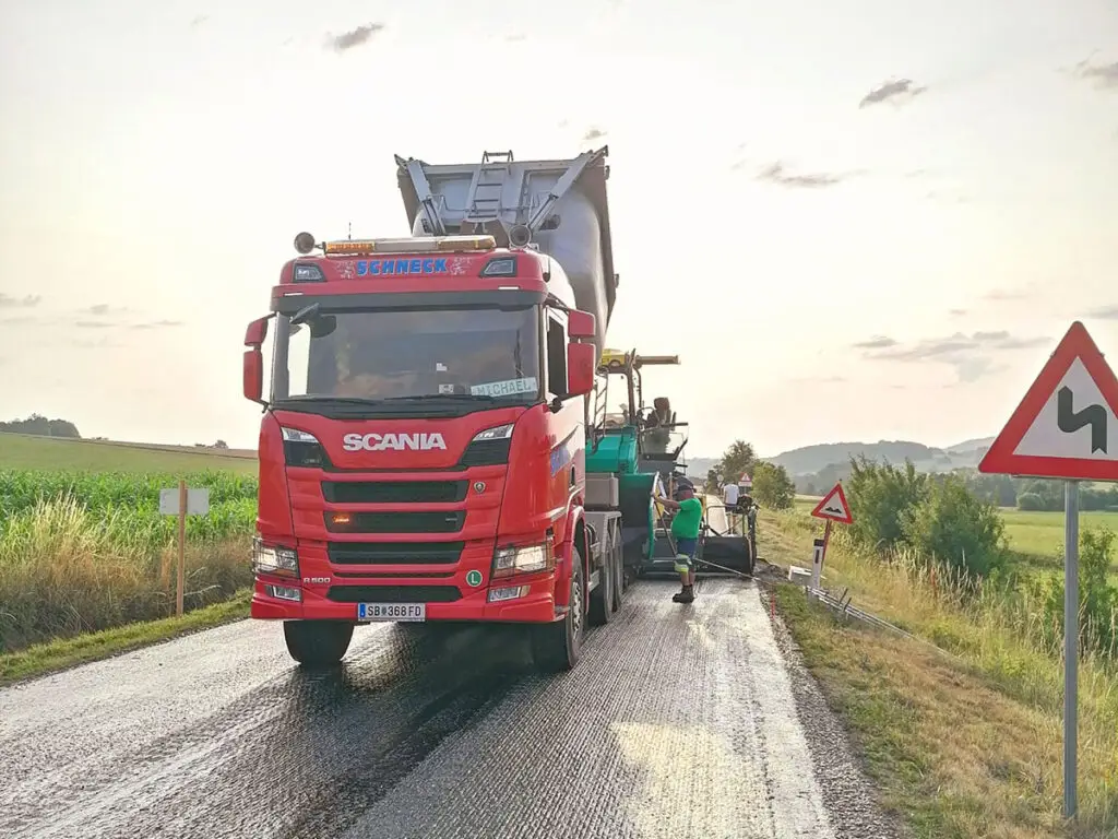 Asphalttransportfahrzeug vor einem Straßenfertiger während Asphaltierungsarbeiten auf einer Landstraße.