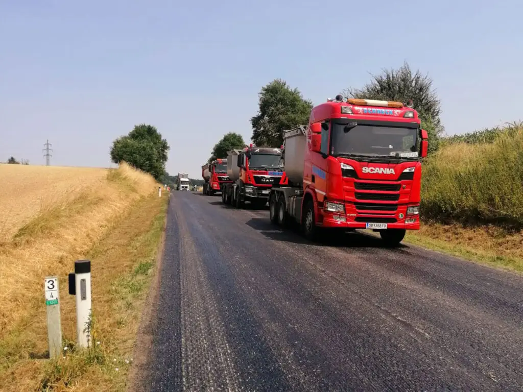 Mehrere Asphalttransporter bei der Materialversorgung einer laufenden Straßenbaustelle.