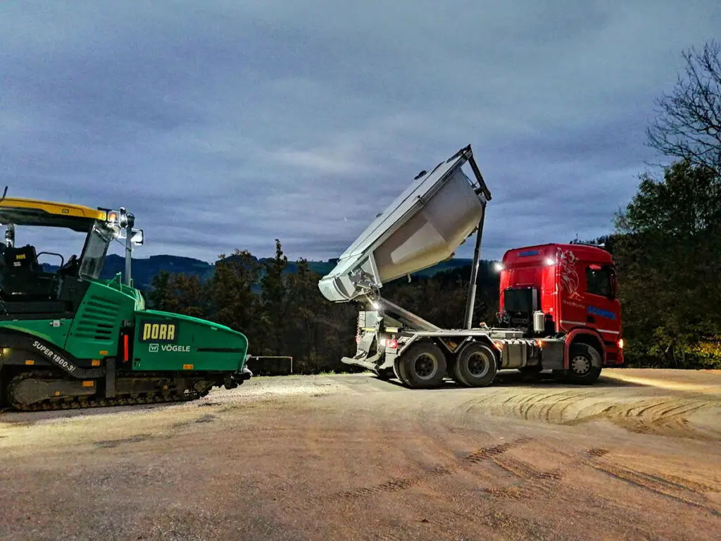 Asphaltmulde beim Entladen von Heißasphalt direkt vor einem Straßenfertiger in den Abendstunden.