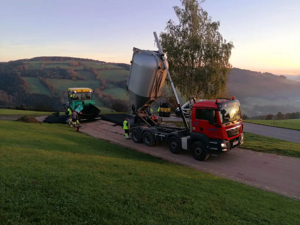 Asphalttransport auf einer ländlichen Straße mit Straßenfertiger und laufenden Asphaltierungsarbeiten im Hintergrund.
