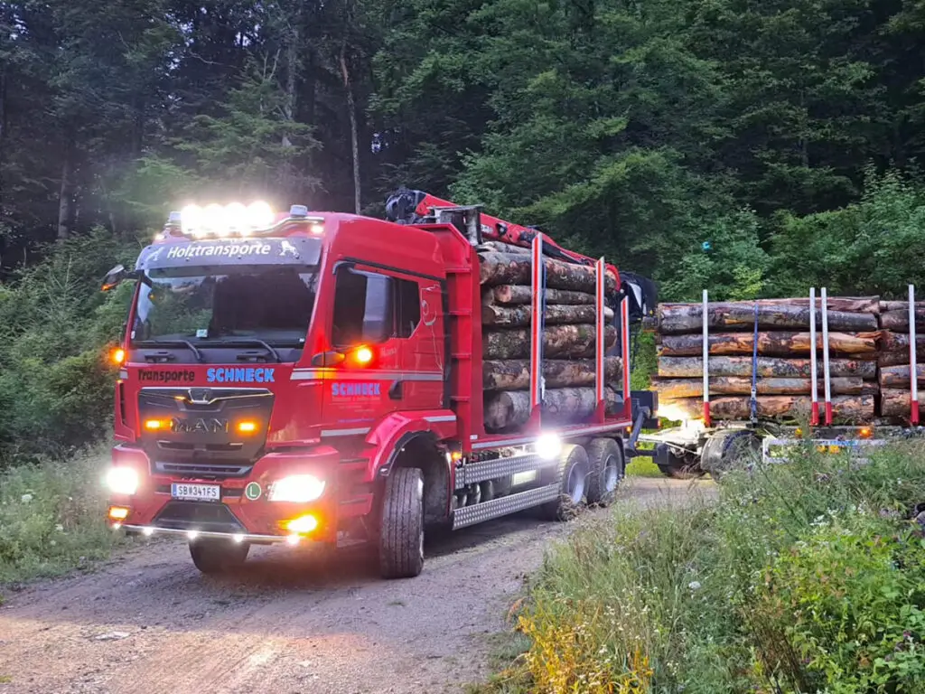 Beladener Rundholz-LKW mit eingeschalteter Beleuchtung auf einer Waldstraße.