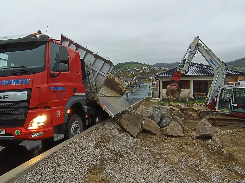 Verladung und Transport von Natursteinen mit LKW und Bagger auf einer Baustelle.