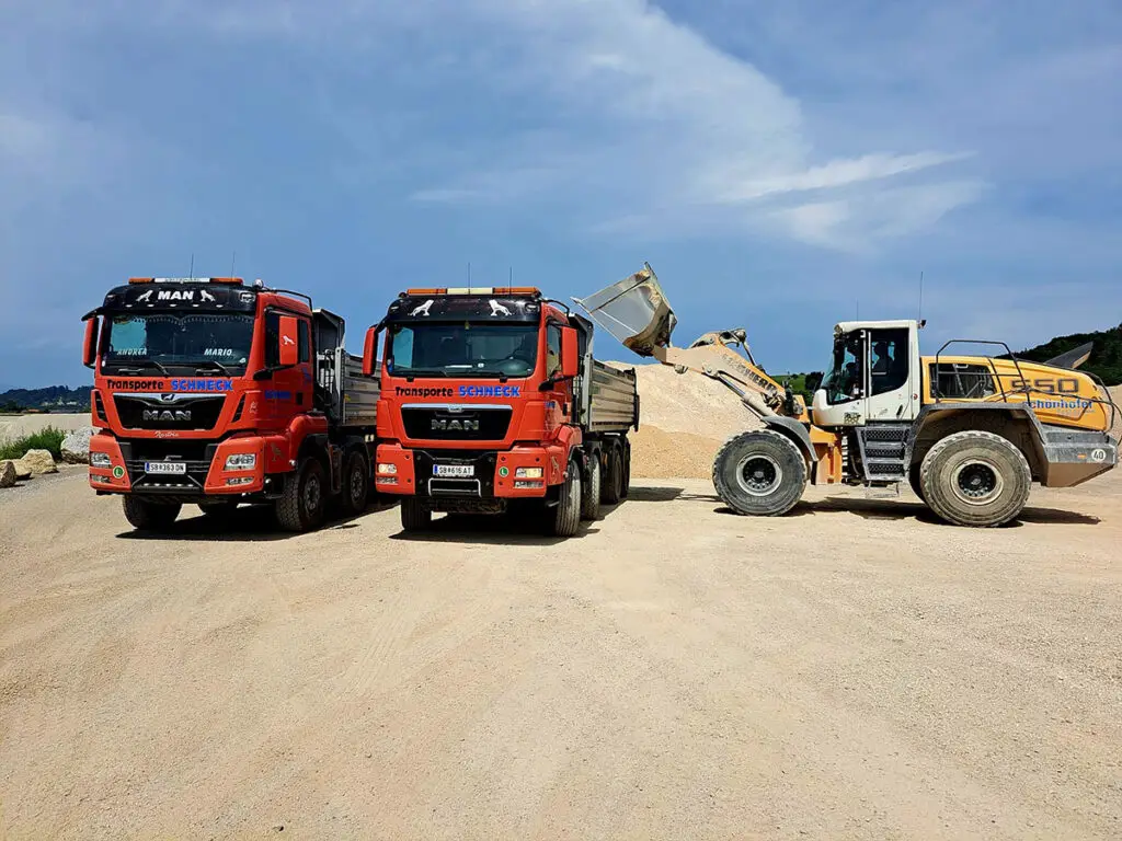 Zwei Schneck-LKW und Radlader beim Verladen von Schotter in einem Materiallager.
