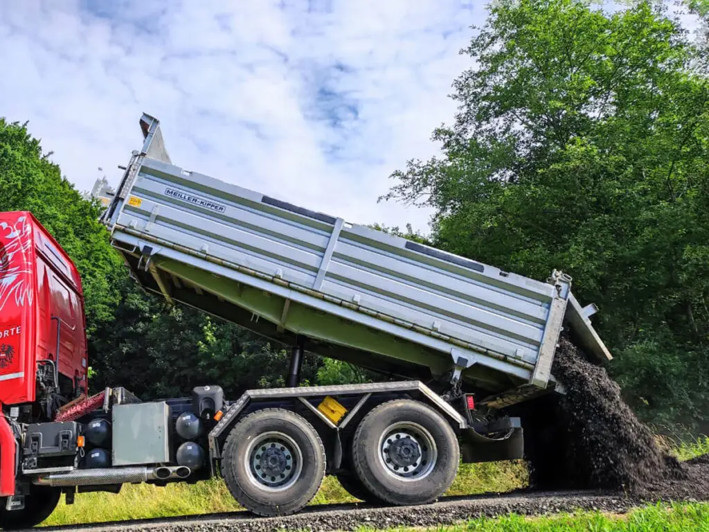 Kipp-LKW beim Abladen von Erd- und Schottermaterial am Straßenrand.