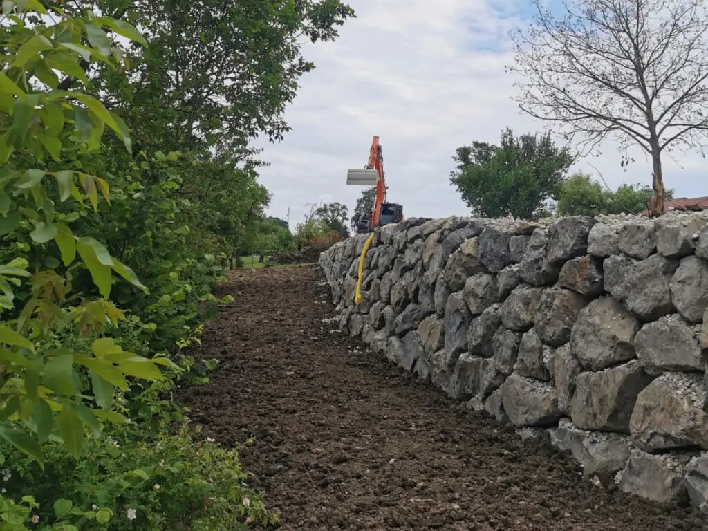 Lange Naturstein-Wurfsteinmauer entlang eines Geländestreifens mit Bagger im Hintergrund.