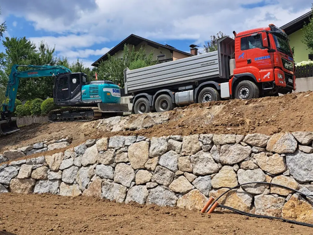 Wurfsteinmauer bei einem Bauprojekt mit LKW und Bagger auf dem darüberliegenden Gelände.