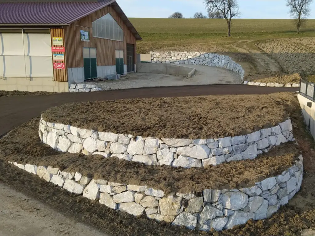 Mehrstufige Wurfsteinmauer zur Geländegestaltung vor einem landwirtschaftlichen Gebäude.