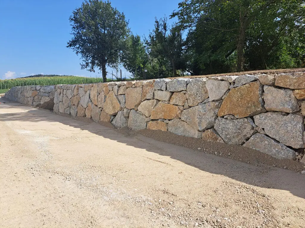 Fertiggestellte Naturstein-Wurfsteinmauer entlang einer Zufahrt im ländlichen Bereich.