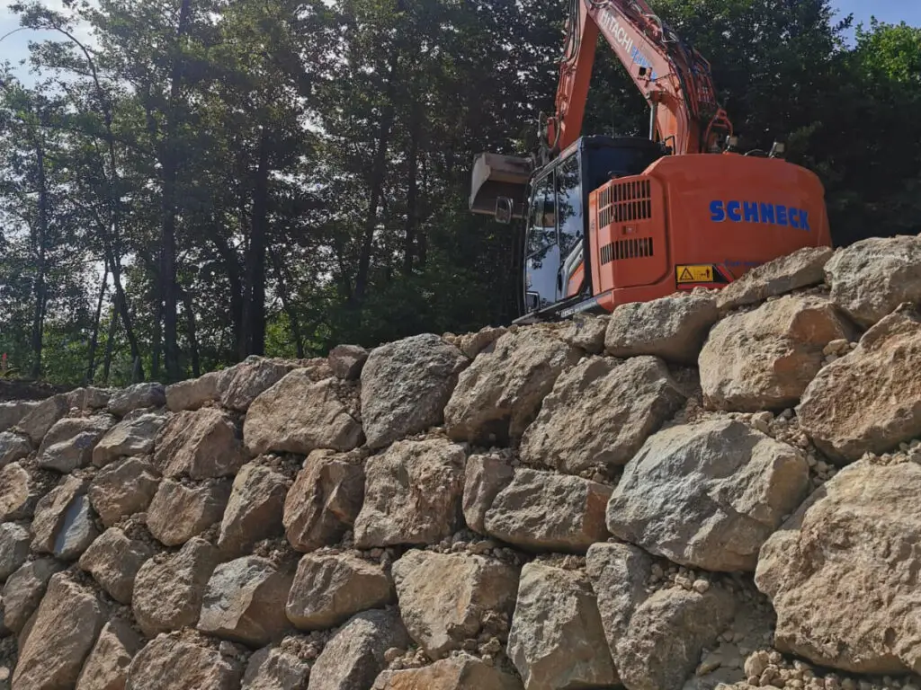 Baggerarbeiten an einer Wurfsteinmauer aus großen Natursteinen zur Hangsicherung.
